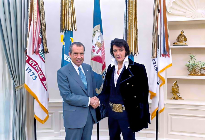 Two men shaking hands in front of state flags and a trophy.