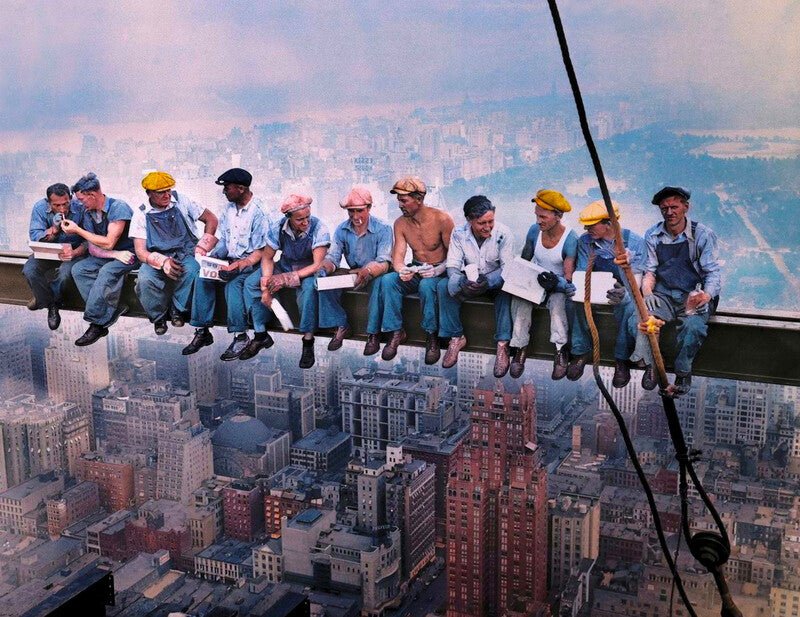 A group of men wearing hard hats are sitting on a metal beam, overlooking a cityscape below.