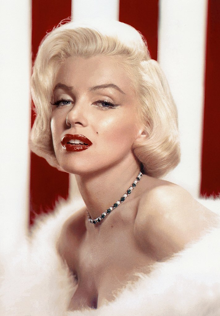 A close-up portrait of a woman with blonde hair and bold red lipstick, wearing a fur coat and a necklace.