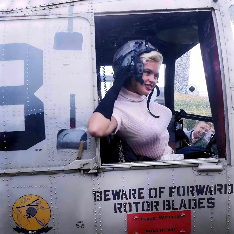 A woman in a pink sweater and helmet is sitting in the cockpit of a helicopter, smiling and looking out the window.