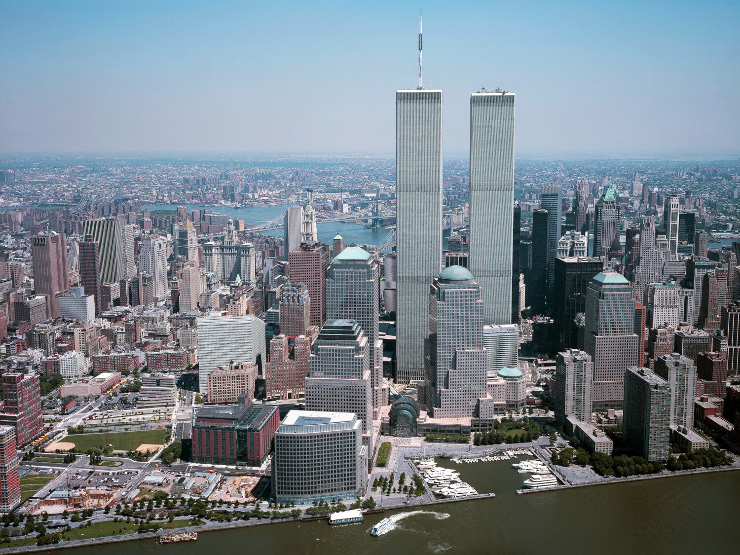 a panoramic view of the New York City skyline, featuring the iconic twin towers of the World Trade Center. The cityscape is surrounded by a body of water, with boats visible in the harbor. The sky is clear and blue, creating a vibrant and bustling urban atmosphere.
