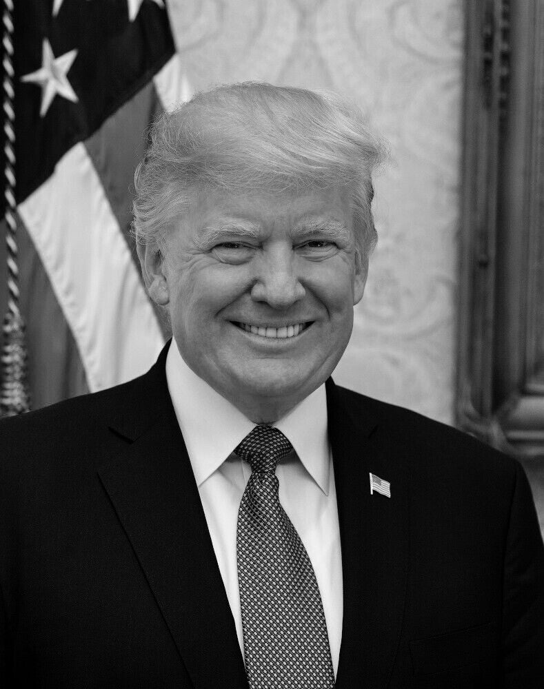 A black and white portrait of a smiling man wearing a suit and tie, standing in front of an American flag.