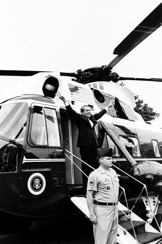 A black and white image showing a man in a suit and tie standing in the doorway of a helicopter, with another man in a military uniform standing nearby.