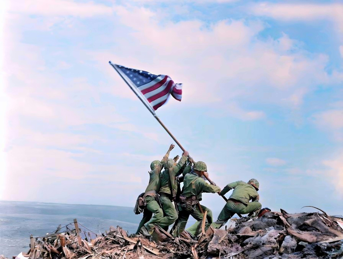 A group of soldiers in green uniforms are raising an American flag on a rocky hill, with the ocean visible in the background.