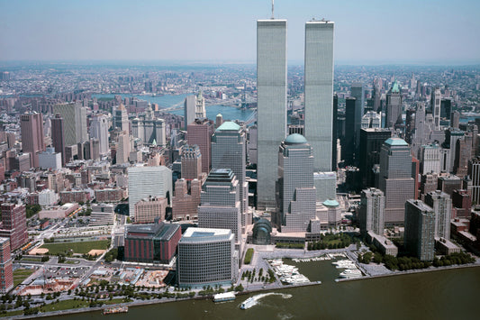 a panoramic view of the New York City skyline, featuring the iconic twin towers of the World Trade Center. The cityscape is densely populated with numerous high-rise buildings, and the waterway in the foreground adds a natural element to the urban landscape.