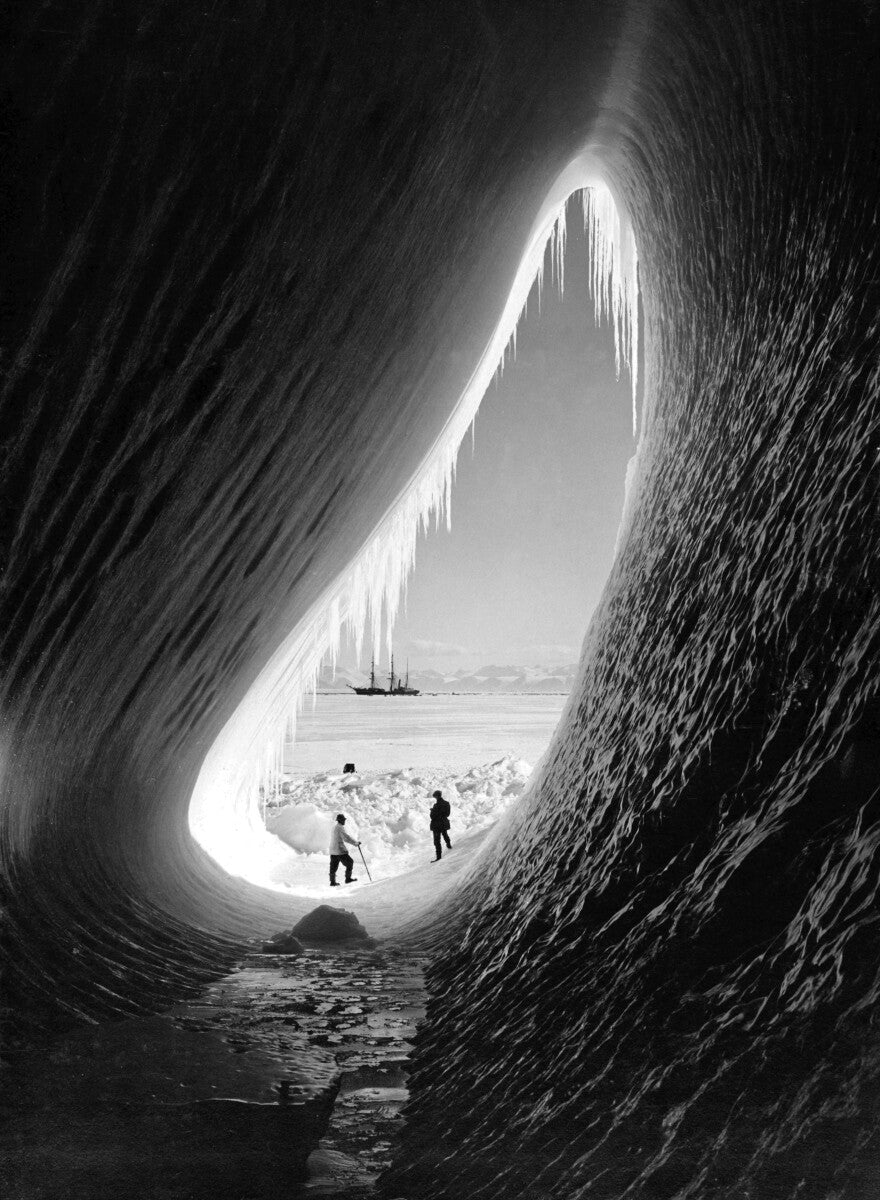 A black and white image depicting a cave with icicles hanging from the ceiling, and two people standing in the doorway of the cave.