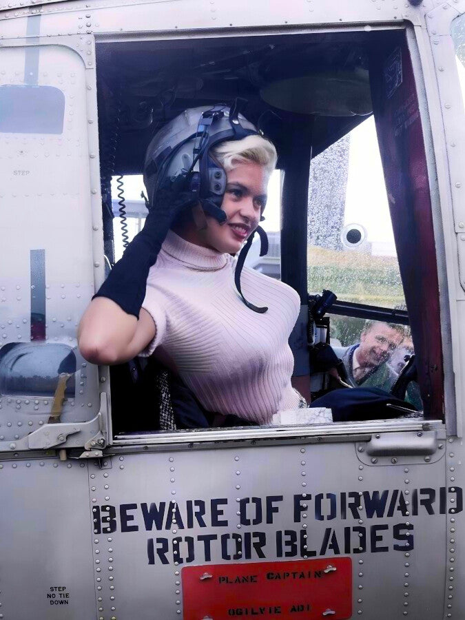 A woman in a pink shirt and helmet is sitting in the cockpit of a helicopter, with a sign on the side of the helicopter that reads "BEWARE OF FORWARD ROTOR BLADES".
